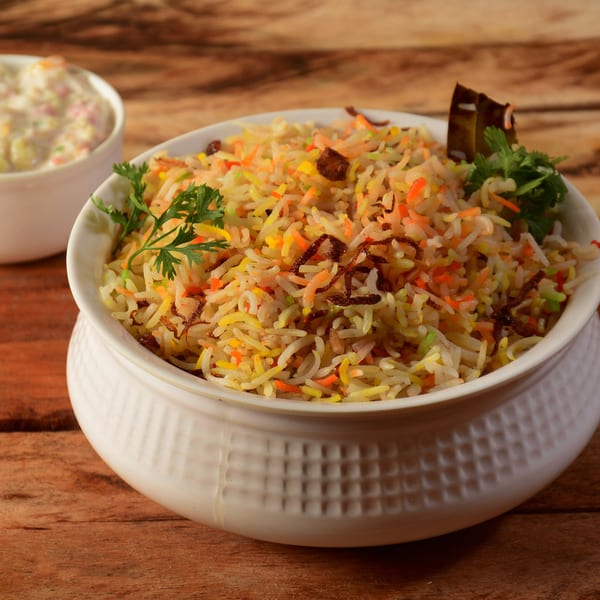 Colorful Indian biryani rice with fried onions and cilantro in a white bowl, with raita in the background.